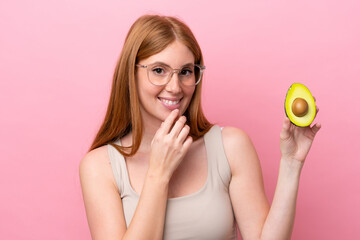 Young redhead woman holding an avocado isolated on pink background looking up while smiling