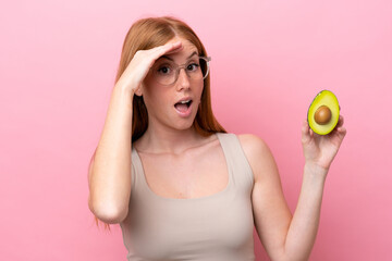 Young redhead woman holding an avocado isolated on pink background doing surprise gesture while...