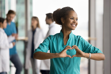 Happy dreamy beautiful African doctor woman promoting cardio healthcare checkup, joining fingers at...