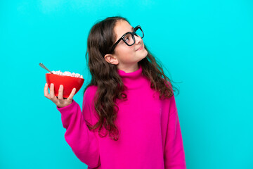 Little girl holding a bowl of cereals isolated on blue background looking up while smiling
