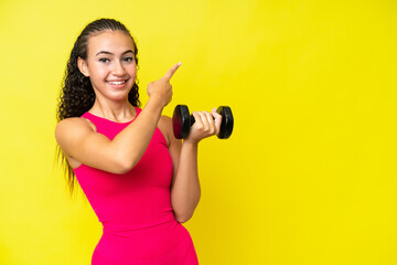 Young sport woman making weightlifting isolated on yellow background pointing back