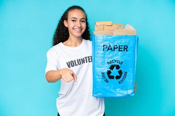 Young woman holding a recycling bag full of paper to recycle isolated on blue background points finger at you with a confident expression