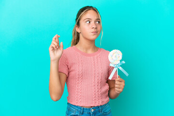 Little Caucasian girl holding a lollipop isolated on blue background with fingers crossing and...