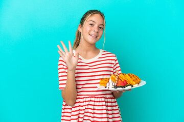 Little caucasian girl holding waffles isolated on blue background saluting with hand with happy expression