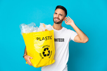 Young caucasian man holding a bag full of plastic bottles to recycle isolated on blue background...