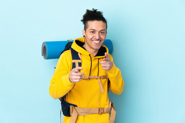 Young mountaineer man with a big backpack over isolated blue background points finger at you