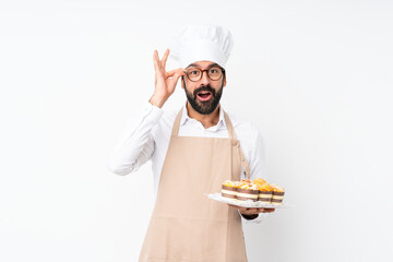 Young man holding muffin cake over isolated white background with glasses and surprised