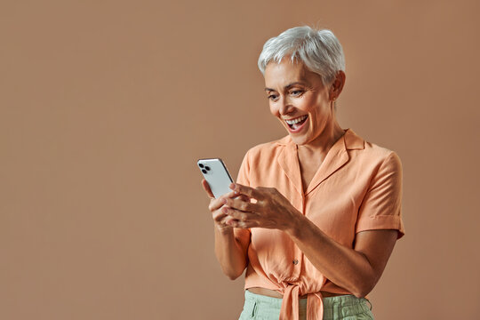 Portrait of a stylish modern beautiful gray haired middle aged woman in a pastel orange shirt and green pants holding a white phone and looking at it with joy and surprise.