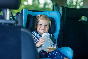 Adorable baby girl with blue eyes sitting in car safety seat. Toddler child going on family vacations and jorney. Smiling happy child during traffic jam, drinking milk from bottle and eating bisquit
