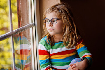 Adorable little preschool girl with eyeglasses sitting by the window. Thoughtful child looking out....