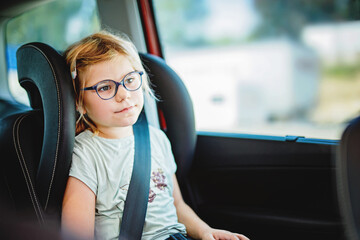 Little Preschool Girl Sitting in Her Car Seat. Happy Child with Eyeglasses looking out of the window. Smiling kid on the Way to Family Vacations during Traffic Jam.