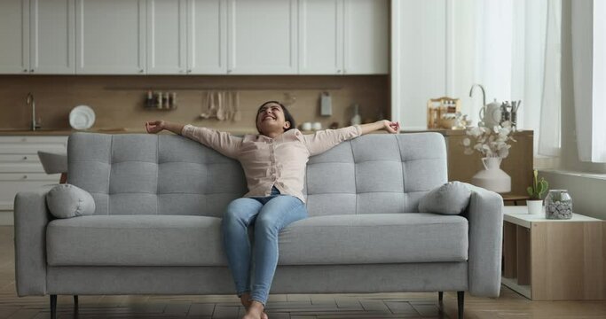 Cheerful excited Indian homeowner walking in cozy living room, falling on comfortable couch, relaxing on sofa with open arms, smiling, laughing, enjoying homey comfort, leisure at home