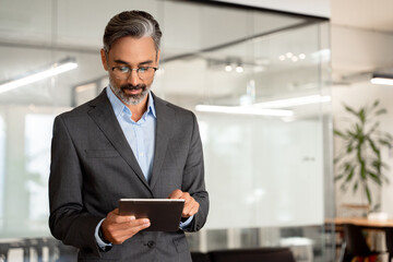 Happy Latin Hispanic bearded stylish mature adult professional business man, smiling Indian senior businessman CEO holding digital tablet using fintech tab application standing inside company office.
