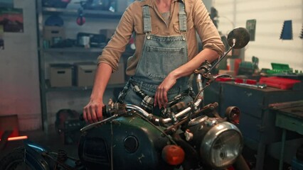 Portrait of young confident woman standing near vintage retro motorcycle and looking directly at camera. Beautiful female auto mechanic working in garage. Woman biker tuning motorcycle in workshop.