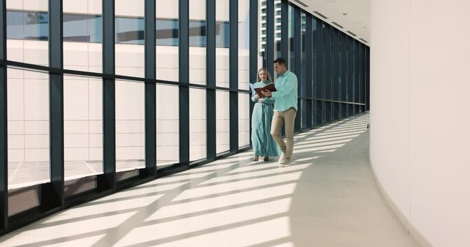 Two young male female colleagues, businesspeople talking, discussing contract details, making last changes before meeting with partners, consider joint work walking in modern office building hallway