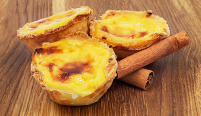 Three Pastel de nata or Portuguese egg tart and cinnamon sticks on a wooden brown background. Pastel de Belm is a small dessert, a cup-shaped pastry made from puff pastry with custard.