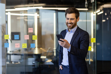 Businessman with phone in hands inside office at workplace, joyful successful man in business suit...