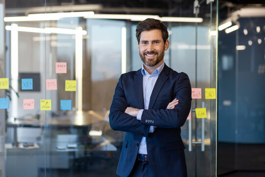 Portrait of successful businessman inside office in business suit, man with crossed arms smiling and looking at camera, experienced financier boss near window, investor.