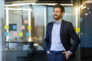 Successful joyful businessman experienced man inside office at workplace, boss in business suit smiling joyfully with hands in pockets, looking satisfied with achievement results out the window. - Powered by Adobe