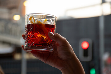 female hand toasting with glass of red negroni drink with orange slices in blurred traffic light