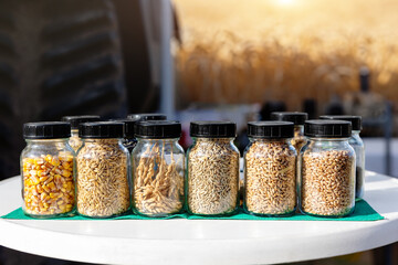 Different kinds of cereal crops seed grain in glass jar row at agricultural expo exhibition. Harvester machine tractor on background. Sowing material sample biotechnology test analysis