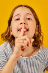 Fototapeta premium Young Girl Gesturing Silence With Finger on Lips Against Bright Yellow Background