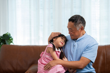 Happy Asian senior grandfather sits on a couch with his granddaughter and plays with his...