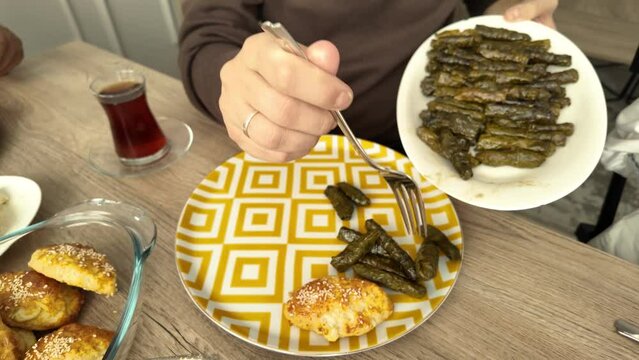 Man takes stuffed grape leaves to her plate on a Turkish breakfast table filled with traditional foods. Family reunion.