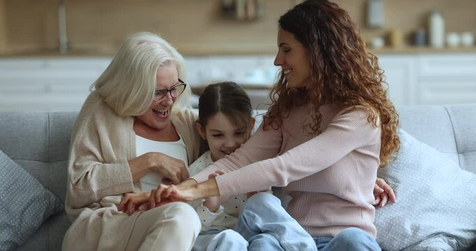 Cheerful grandma and mother tickling playful little kid girl on home sofa, playing with child, laughing, shouting, having fun, enjoying playtime, leisure together, hugging with love, care