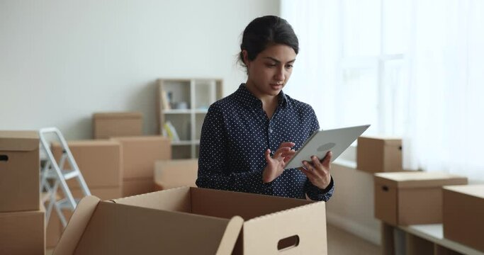 Serious Internet shop manager packing parcel for customer in e-commerce storage, checking open cardboard box, using tablet computer, processing online order, preparing package for delivery