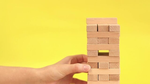 A woman plays Jenga on a yellow background. The concept of family vacation, holidays, entertainment. A woman's hand takes out a block from a wooden toy tower. A game