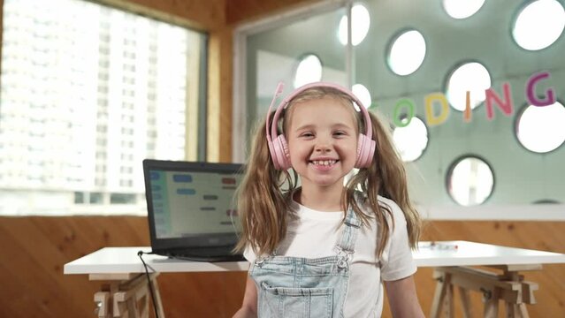 Smiling girl using laptop and turn around to show thumb up to camera. Cute child wearing headphone while working by using laptop writing code in STEM technology education. Online learning. Erudition.