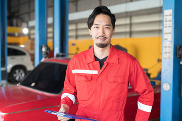 Portrait of Asian male mechanic working, standing and holding clipboard, wearing red uniform in...