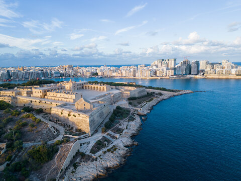 A tourist attraction Fort Manoel at sunset with Silema skyline. Low altitude aerial view on fort at golden hour. Natural blue and golden colors with clouds in the sky.