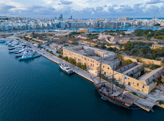 Naklejka premium Beautiful sunset on Lazaretto of Manoel Island with Yacht Marina and old wooden sailing ship with French flag. Aerial view on a city skyline with natural golden and blue colors. Clouds in the sky.