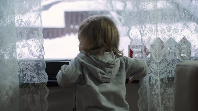 A child is standing in front of a window, looking outside at the first snow and the snowy yard.