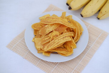 Crispy banana chips on a white plate with fresh bananas.