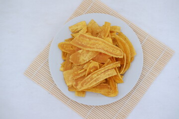 Crispy banana chips on a white plate with fresh bananas.