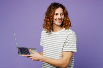 Sideways young IT man he wearing grey striped t-shirt casual clothes hold use work on laptop pc computer look aside on area isolated on plain pastel light purple background studio. Lifestyle concept.
