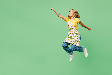 Full body side view young housewife housekeeper chef cook baker woman wear apron yellow t-shirt jump high do super hero gesture pov fly isolated on plain green background studio. Cooking food concept. - obrazy, fototapety, plakaty