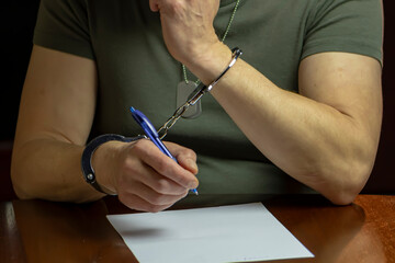 A handcuffed military man signs a document on white paper, interrogation and testimony. - obrazy, fototapety, plakaty