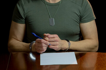 A handcuffed man sitting at a wooden table, a blank sheet of paper in front of him and a pen in his...