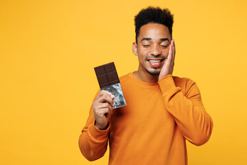 Young man wear orange sweatshirt casual clothes hold eat bar of chocolate put hand on face close...