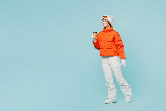 Full body side view young skier woman wears warm padded windbreaker jacket hat ski goggles mask drink cup coffee travel rest spend weekend winter season in mountains isolated on plain blue background.