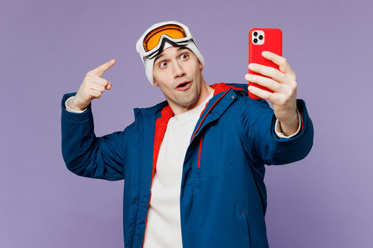 Skier man wear warm blue windbreaker jacket point finger on ski goggles mask hat do selfie shot mobile cell phone spend extreme weekend winter season in mountains isolated on plain purple background.