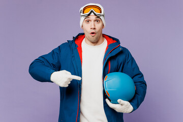 Skier shocked amazed man wear warm blue windbreaker jacket ski goggles mask hat hold point index finger on hardhat spend extreme weekend winter season in mountains isolated on plain purple background.