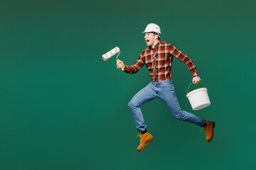 Full body young employee laborer man wearing red shirt hardhat jump high hold paint roller paint tin isolated on plain green background. Instruments for renovation apartment room. Repair home concept.