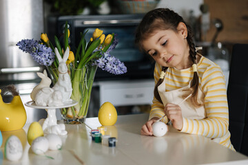 Adorable little girl painting eggs on kitchen at home. Cute girl in apron. Background of easter...