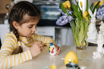 Fototapeta na wymiar Cute four year girl in beige apron sitting on kitchen table and painting easter eggs