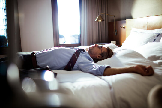 Middle aged businessman lying down and resting on a bed in a hotel room after a long day of traveling and working